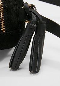 Sorte læderkvaster fastgjort til en guldring på en sort ruskinds taske nær en guldlås.