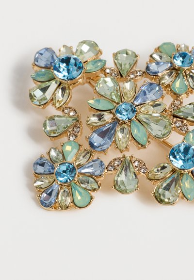 Καρφίτσα με λουλουδάτο σχέδιο σε χρυσό τόνο με μπλε και πράσινες σταγόνες και στρογγυλές γυαλιστερές πολύτιμες πέτρες τοποθετημένες σε στρωμένα πέταλα.
