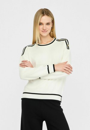 Pull en maille blanche avec des accents rayés noirs aux poignets et au col. Présente des boutons décoratifs aux épaules. Texture lisse.