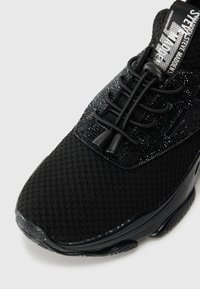 Chaussure de sport noire avec une tige en tissu texturé, des lacets élastiques sans nœud et une languette de marque affichant le texte "shadow" en blanc.