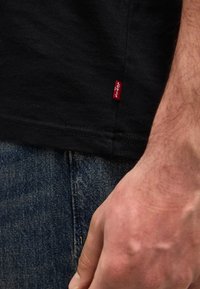 Κοντινό πλάνο ενός ατόμου που φορά μπλε τζιν και μαύρο πουκάμισο με μια μικρή κόκκινη ετικέτα Levi's, το χέρι ακουμπάει στο πλάι.