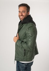 Giacca di pelle verde con zip frontale, cappuccio nero e tasche laterali. Presenta un materiale testurizzato e un design aderente con dettagli sulle spalle.
