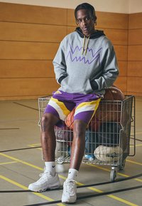 Sudadera gris con gráfico morado, forro interior negro, cordón amarillo; pantalones cortos con patrón morado y blanco; zapatillas blancas; cesta de alambre con pelotas deportivas.