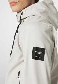 Light gray hooded jacket made of smooth fabric, featuring a black patch with geographic coordinates on the sleeve and adjustable drawstrings.