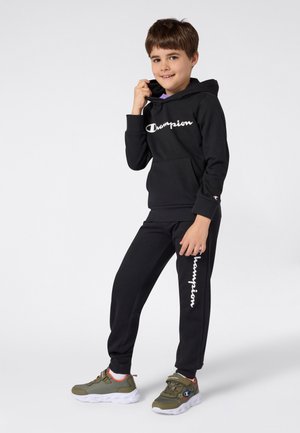 Conjunto de sudadera negra y pantalones con el logo blanco de "Champion". La sudadera tiene capucha y un bolsillo frontal; los pantalones llevan el logo a lo largo de la pierna. Los zapatos son verdes con detalles.