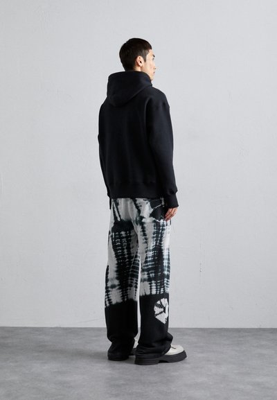 Schwarzer Kapuzenpullover mit gerippten Bündchen; kombiniert mit locker sitzenden, weiß-schwarzen Tie-Dye-Hosen mit auffälligem Muster und seitlichen Taschen.