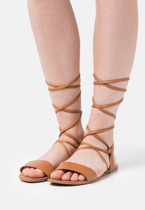 Sandalias de cuero marrón con suela plana, punta abierta y correas cruzadas que se envuelven alrededor de los tobillos. Diseño sencillo con textura suave.