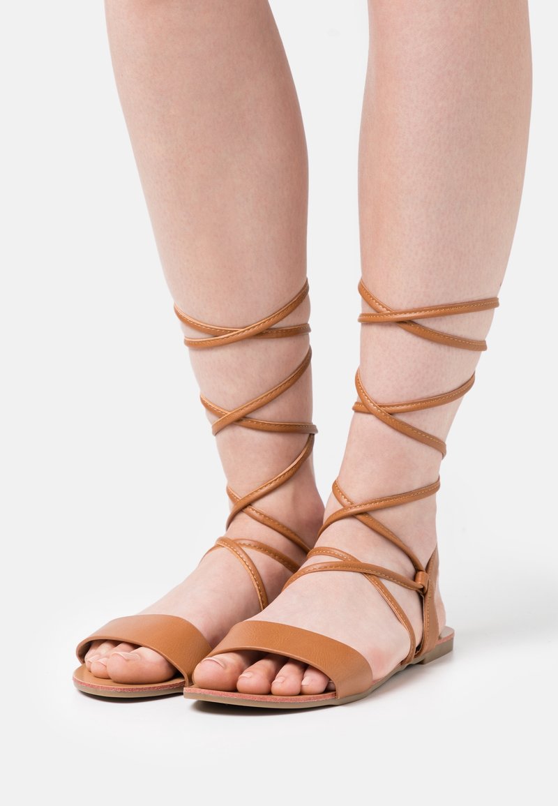 Sandales en cuir marron avec semelle plate, bout ouvert et brides croisées entourant les chevilles. Design simple avec une texture lisse.