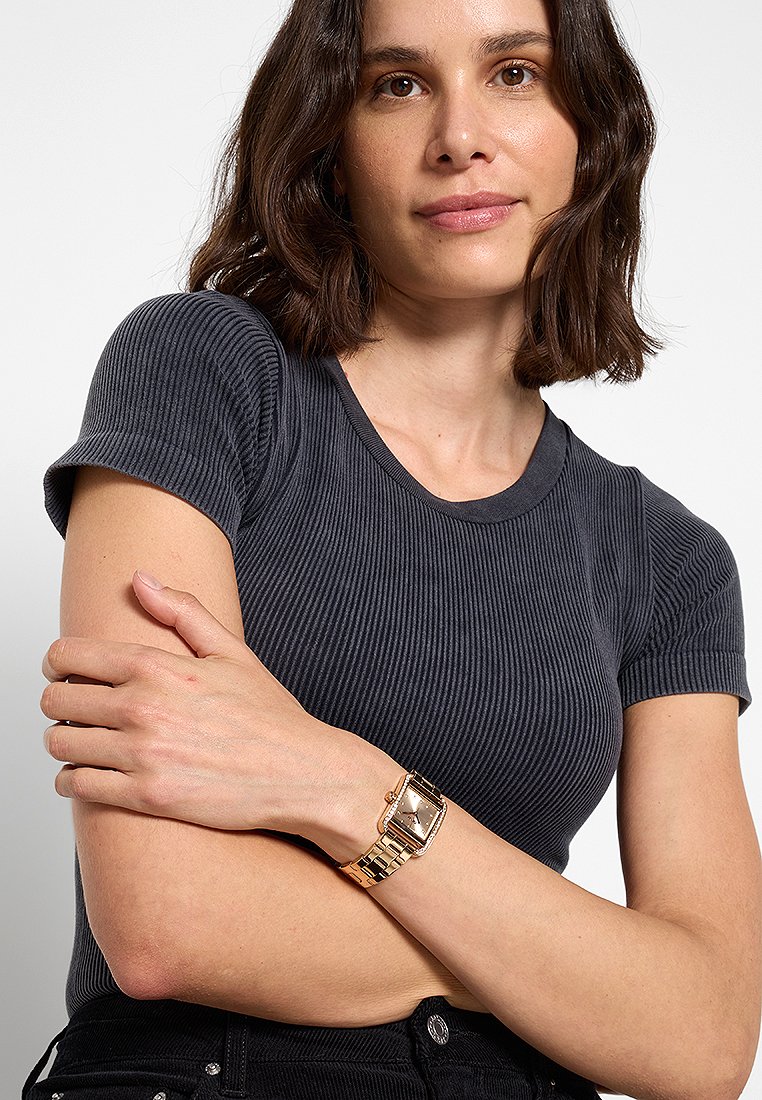 Reloj dorado con esfera cuadrada y pulsera de eslabones. Modelo vistiendo una camiseta oscura de canalé, con los brazos cruzados y el cabello a la altura de los hombros.