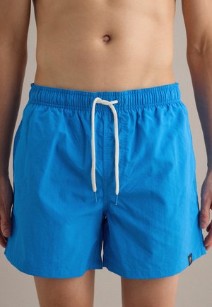 Man wearing mid-thigh bright blue swim trunks with white drawstring and small black logo tag on the left leg.