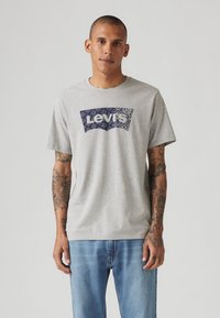 Pilkas medvilnės marškinėlis su tamsiai mėlynu Levi's logotipu su gėlių raštu. Trumpomis rankovėmis, apykakle, laisvu kirpimu, derinamas su džinsais.