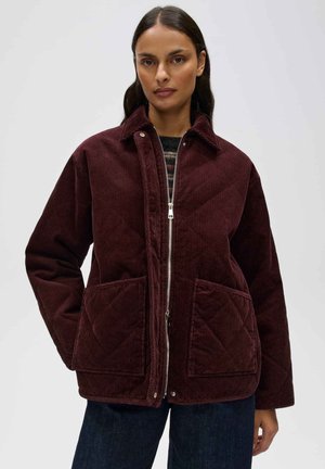 Donna con lunghi capelli scuri indossa una giacca trapuntata in velluto a coste color bordeaux con grandi tasche frontali e jeans scuri su uno sfondo semplice.