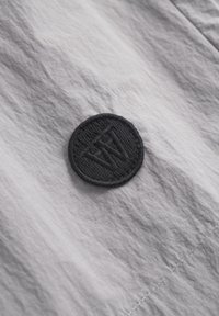 Svijetlosivi tekstil s kružnim crnim izveznim obrascem koji sadrži inicijale. Glatka površina s minimalnim naborima i suptilnim sjajem.