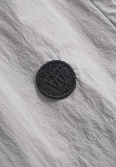 Lys grå stoftekstur med en cirkulær sort broderet lap, der har initialer. Glat overflade med minimal rynkning og subtil glans.