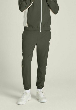 Mann trägt dunkelgrüne Trainingshose und passende Jacke mit weißen Seitenstreifen, weiße Socken und weiße Turnschuhe, steht.