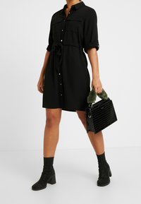 Zwart button-up jurk met korte mouwen, strik in de taille en twee borstzakken. Gecombineerd met een zwarte handtas van krokodillenleer en enkellaarsjes.