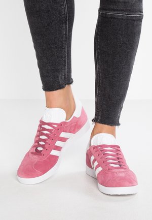 Pieds portant des baskets Adidas en daim rose avec des bandes blanches et des semelles blanches, associées à un jean skinny noir déchiré.