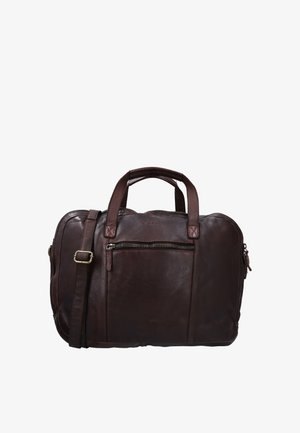 Homme tenant une mallette en cuir marron foncé par les poignées, portant un pantalon beige et une chemise taupe, sur un fond uni.