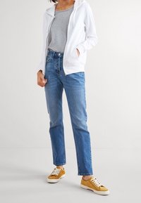Felpa bianca con zip, t-shirt grigia, jeans dritti azzurri chiari e sneakers beige con suole e lacci bianchi. Abbigliamento casual con una vestibilità rilassata.