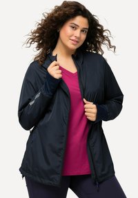Granatowa kurtka zapinana na zamek, z długimi rękawami i mankietami, w połączeniu z różową koszulą z dekoltem w serek. Kurtka ma gładką teksturę i subtelne detale wzornicze.