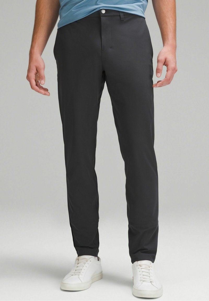 Homme portant un pantalon noir ajusté et des baskets blanches, debout devant un fond uni gris clair.