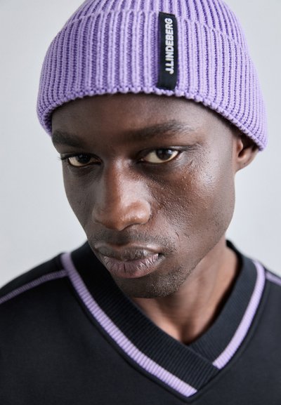 Lilla ribbet strikhue med et mærke; model iført en sort V-hals sweater med lilla nuancer. Nærbillede.