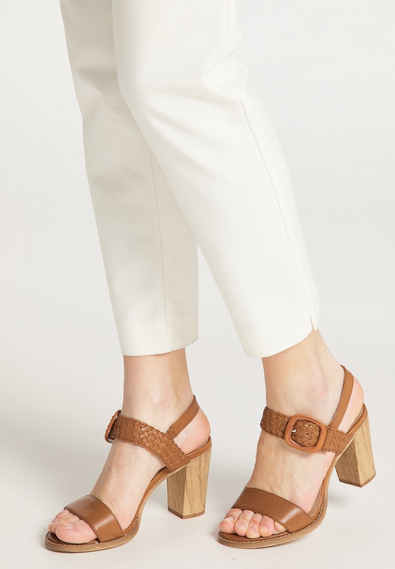 Piedi che indossano sandali beige in pelle con tacco bloccato, dotati di cinghie intrecciate e fibbie, abbinati a pantaloni bianchi a 3/4 su uno sfondo neutro.