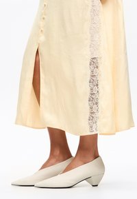 Vestido amarillo claro con detalle de encaje en el costado y una abertura lateral, combinado con tacones beige de punta con un tacón bajo en forma de cono.