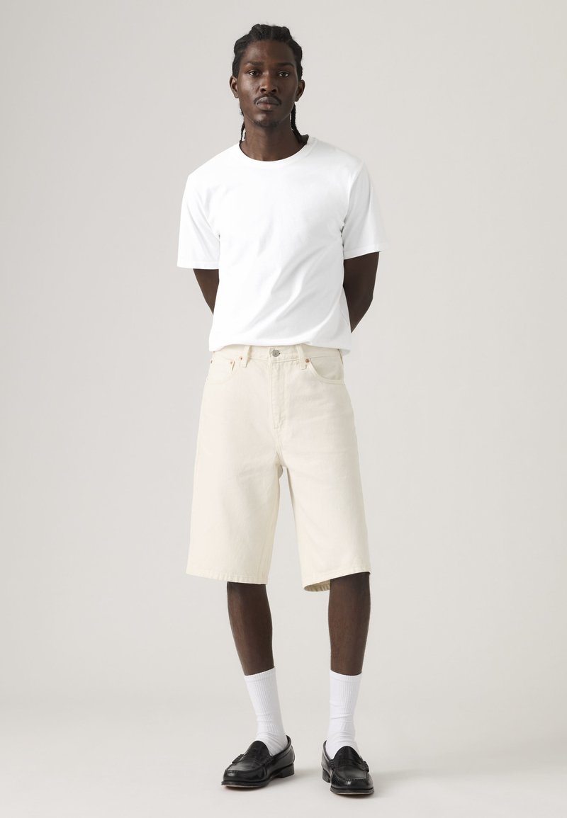 Jeune homme debout, les mains dans le dos, portant un t-shirt blanc, un short beige jusqu'aux genoux, des chaussettes blanches et des chaussures de ville noires, sur un fond uni.