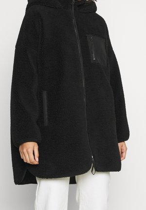 Veste zippée en polaire noire et duveteuse avec capuche, poche zippée sur la poitrine, poches latérales, portée sur un pantalon blanc.