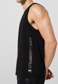 Juodas be rankovių marškinėlis, pagamintas iš minkštos medžiagos, su vertikaliu baltu "KARL LAGERFELD" logotipu šone ir apvalia iškirpte.