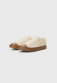 Beige kanvas joggesko med en brun gummitåhette og såle, med en sølvstjerne-logo på siden og hvite snørebånd.