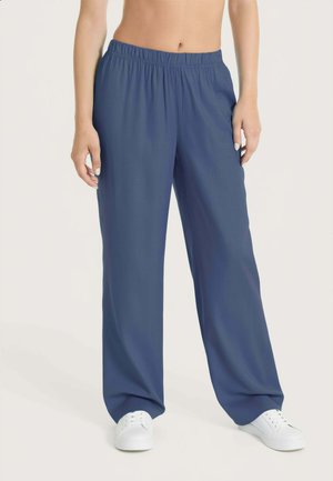 Donna che indossa pantaloni blu larghi con vita elastica e sneakers bianche, in piedi davanti a uno sfondo semplice.