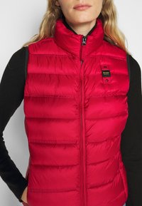 Chaleco acolchado rojo con cuello alto, secciones acolchadas horizontales y cierre de cremallera. Presenta una pequeña etiqueta de marca negra en el lado izquierdo del pecho.