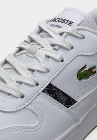 Λευκό αθλητικό παπούτσι Lacoste με λευκά κορδόνια, πράσινο λογότυπο κροκοδείλου και γεωμετρική ριγέ μαύρου σχεδίου στη πλευρά.
