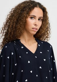 Chemisier bleu marine avec des motifs de cœurs blancs, décolleté en V, manches fluides, texture douce, tissu léger. Design et détails minimalistes.