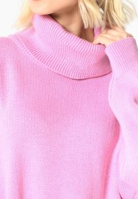 Světle růžový pletený svetr s vysokým žebrovaným límcem a měkkou texturou. Má volné rukávy a uvolněný střih.