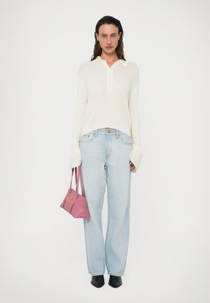Kvinna som står klädd i en krämfärgad ribbad tröja, ljusblå jeans, svarta spetsiga skor, och håller en rosa handväska med guldtryck.