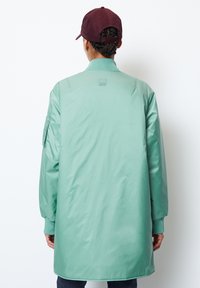 Dlouhá světle zelená bunda s hladkou texturou, žebrovanými manžetami a malou nášivkou na zádech. Doplněná tmavě vínovou čepicí.