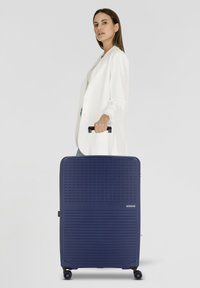 Valigia rigida blu navy con superficie testurizzata, manico telescopico e quattro ruote, posizionata accanto a una persona in giacca bianca.