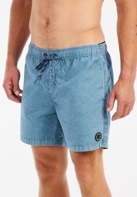 Shorts de bain bleus avec une taille élastique, fermeture à cordon de serrage et petit patch logo rond sur le côté. Tissu doux et texturé.