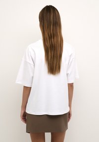 Tricou alb din bumbac, cu mânecă scurtă, croială lejeră și guler rotund. Purtați cu o fustă maro, care prezintă o textură netedă și un design casual.