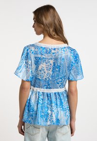 Blusa blu a motivo con bordo in pizzo, maniche corte a volant e design a peplum svasato. Tessuto trasparente con motivi floreali intricati.