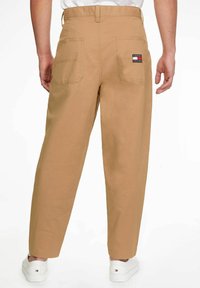 Pantalones de algodón beige con corte relajado, dos bolsillos traseros y un parche con el logotipo de la marca. Los puños están ligeramente ajustados y la tela parece suave.