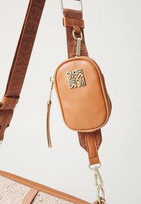 Sac en cuir tan avec un logo doré, comprenant une petite pochette amovible. Sangle marron avec une texture contrastante et des accents en métal.