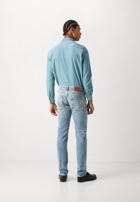 Camisa de manga larga azul clara con cuello recto, combinada con jeans azul claro deslavados. Zapatos negros y una etiqueta de cuero en la cintura son visibles.