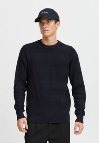 Marineblå strikket genser med en teksturert fiskebeinmønster, rund hals og lange ermer. Kombinert med en matching lue med tekst.