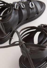 Sandales en cuir marron foncé avec plusieurs brides et fermetures à boucle, ornées de lacets noués à la cheville sur une surface claire.