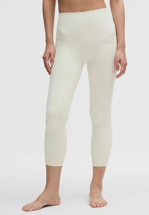 Mujer con leggings cortos de cintura alta y color crema, de pie descalza frente a un fondo claro y liso.