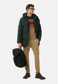 Padded green jacket, wzorzysty sweter w beżowo-czerwonym kolorze, khaki spodnie cargo, czarny plecak i ciemnozielone buty turystyczne.